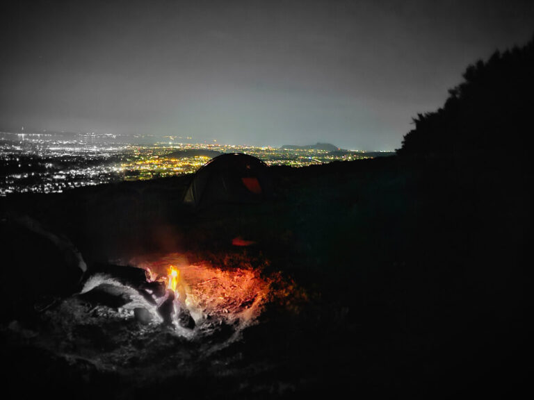 A tent and campfire at night above city lights
